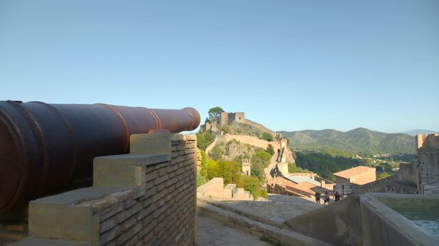 Historic fortress terrace with old cannon overlooking castle walls, terracotta rooftops, green valley, and distant mountains under clear blue sky