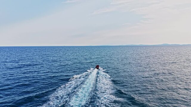 A small boat moves swiftly over the ocean waves, leaving a white wake behind. The sea stretches far into the horizon. The sky is mostly clear with some clouds.