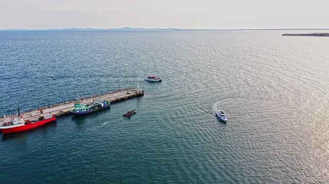 Several boats are maneuvering near a pier with people visible on the dock. The water appears calm under clear skies and distant land is seen on the horizon.