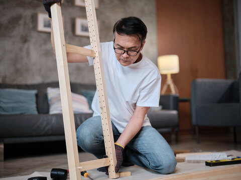 Asian mature man self-assembling flat-pack furniture with DIY tools, checking accuracy and strength of wooden parts, home improvement, craft installation, hobby workshop, handyman active lifestyle.