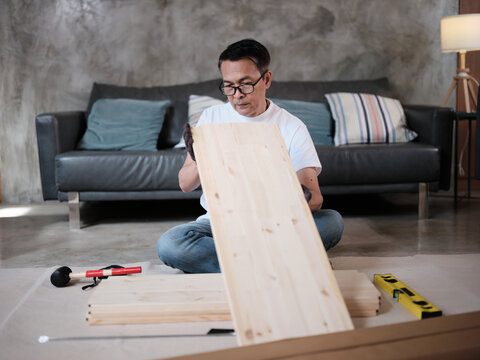 Asian mature man preparing flat-pack furniture parts, checking wooden board parts, self-assembly with carpentry diy tools, home improvement, craft install hobby workshop, handyman active lifestyle.
