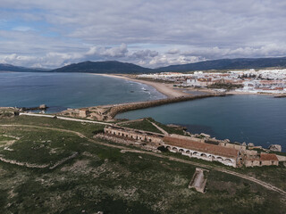 Aerial view of Isla de las Palomas, ancient stone fortress ruins with arched walls overlooking a coastal town, sandy beach and harbor breakwater beneath dramatic cloudy sky in Tarifa, Spain © Felix Morion