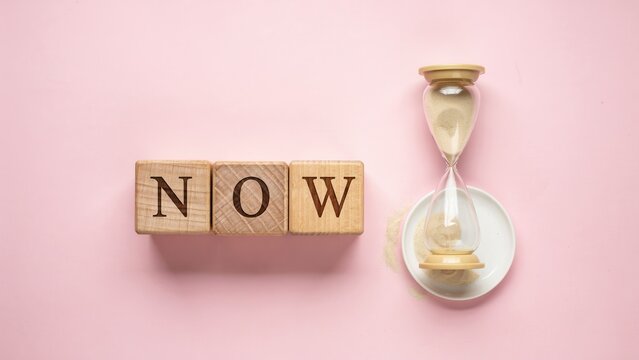 Urgency concept with NOW word on wooden blocks, hourglass with running sand on pink background, time management and deadline metaphor