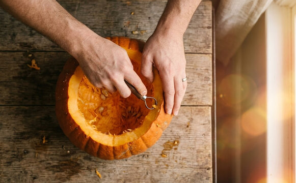 Top view of a man scraping out seeds from a fresh pumpkin for Halloween carving on a wooden table