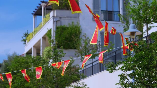 A string of red and yellow flags of Montenegro flutters in the wind against a white multistory building with balconies and green trees, decorating an outdoor space for a celebration or diplomatic.