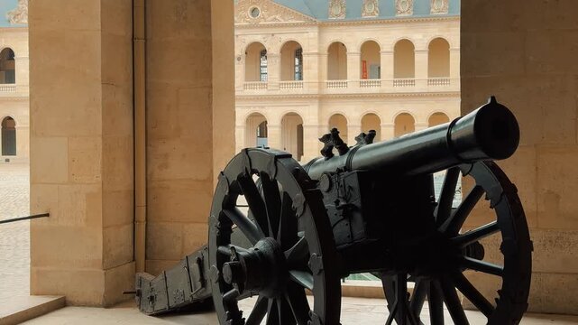 A vintage military cannon stands inside an arched gallery overlooking the grand courtyard and classical architecture of Les Invalides museum in Paris, France, on a cloudy daylight afternoon.

