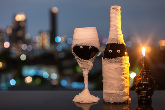 Halloween mummy wine bottle and glass with a lit skull candle against a city skyline at night.