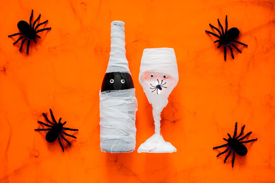Halloween mummy wine bottle and glass wrapped in gauze with googly eyes and spiders on orange background.