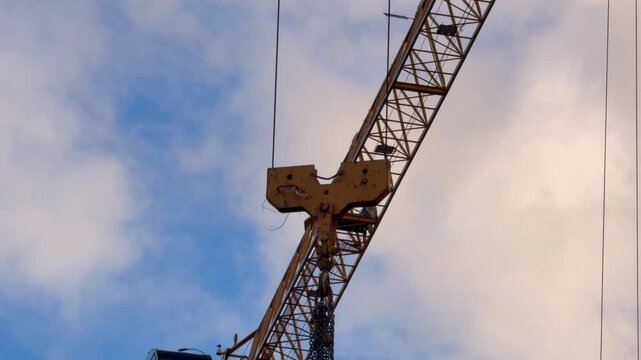A yellow construction crane boom with pulley hook and hanging chains shifts position against a partly cloudy sky, showing heavy machinery in operation at a building site.