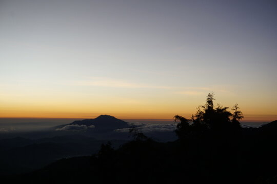 Dramatic mountain silhouette at sunrise with orange and blue sky over misty clouds and pine trees in the foreground