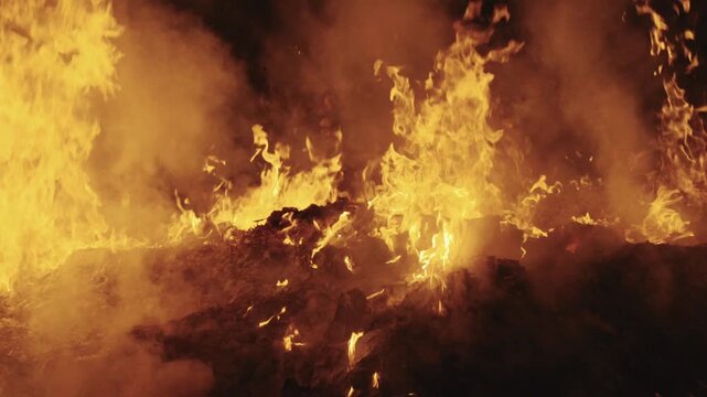 Medium static shot capturing vivid yellow-orange flames actively leaping across an extensive pile of burning ceremonial paper offerings with thick plumes of smoke drifting through the blaze.