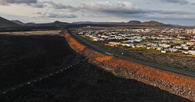 Wide drone view of Grindavik, Iceland, with a long lava defense berm separating the residential town from a vast black lava field beneath rugged mountains and cloudy skies.