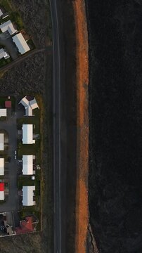 Vertical top-down drone view of Grindavik, Iceland, showing homes beside a paved road and long orange defense berm separating the neighborhood from a dark lava field.
