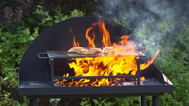 Slices of bread toast on a metal griddle above a wood fire as orange flames rise and smoke drifts through a garden barbecue setting.