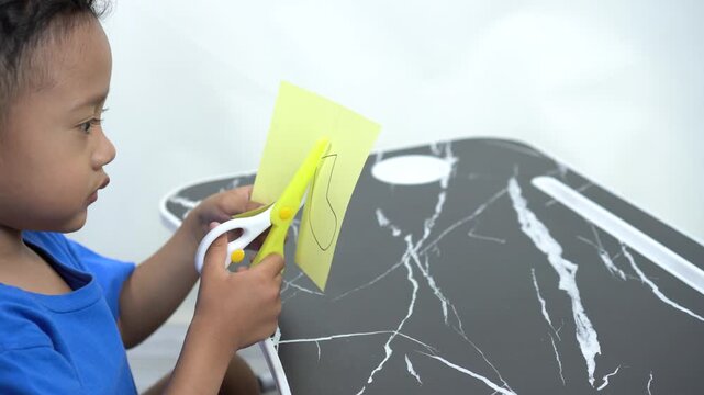 A handsome Asian preschool-aged boy is concentrating on cutting out a pattern from yellow paper to practice his motor skills, using child-friendly yellow-and-white scissors.