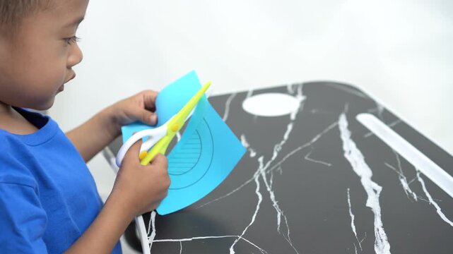 A handsome Asian preschool-aged boy is concentrating on cutting out a pattern from blue paper to practice his motor skills, using child-friendly yellow-and-white scissors.