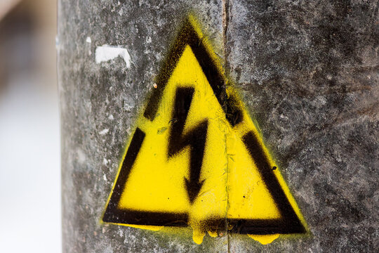 Bright yellow high voltage warning sign spray painted on textured concrete lighting pole outdoors, sharp symbol detail