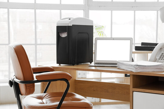 Modern shredder with documents and blank laptop on desk in office