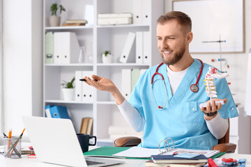 Handsome medical intern with spine model video chatting at table in clinic