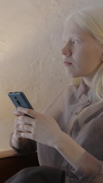 Attractive albino girl messaging with smart phone holding modern gadget sitting in leather sofa in stylish room at home. Communication and people concept.