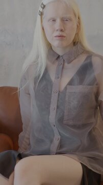 Slow motion portrait of beautiful albino woman in stylish clothing sitting on couch in luxury apartment looking at camera. People and interior concept.