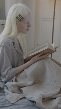 Slow motion of beautiful albino lady in pajamas reading interesting book in bed in luxury bedroom at home. Literature and people concept.