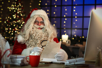 Fototapeta premium Santa Claus with magnifier reading letter at home on Christmas eve