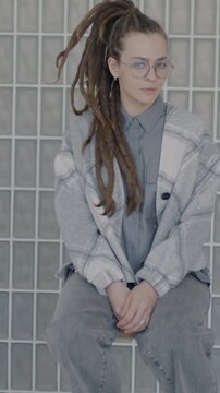 Slow motion portrait of young woman with dreadlocks wearing stylish outfit sitting indoors in creative studio looking at camera. People and individuality concept.