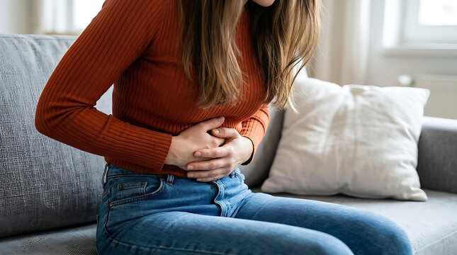 Young Woman Suffering from Stomach Pain While Sitting on Sofa at Home, Abdominal Cramps and Digestive Health Concept