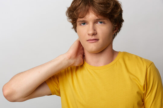 Young man with curly hair in a yellow tshirt looks directly at camera, one hand on the back of his neck. Studio portrait on gray background. Concept of ease and vulnerability.