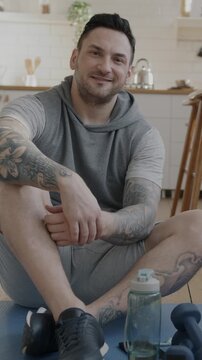 Slow motion portrait of attractive young sportsman smiling looking at camera sitting on yoga mat in apartment. Active lifestyle and sports concept.