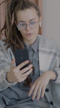 Beautiful young lady with dreadlocks making online video call with smartphone and wireless earphones speaking in luxury apartment. Communication and people concept.