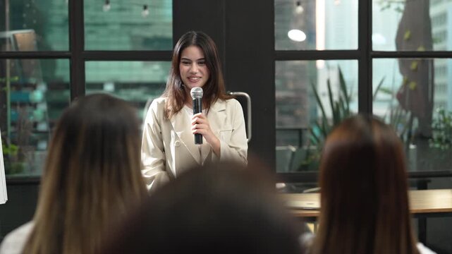Young businesswoman giving presentation with microphone to colleagues in modern office, speaking during corporate training, team meeting and professional workplace communication event.