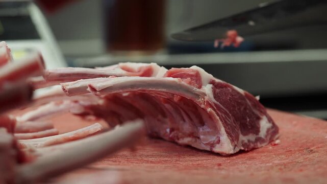 A chef cuts raw lamb ribs with a sharp knife on a red cutting board in a professional kitchen.