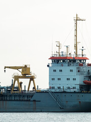 Large dredger ship with yellow deck crane, pipes and white bridge with orange roof anchored on calm river, rusty steel hull and industrial marine equipment under hazy sky © Tavan