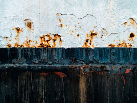Rusty steel ship hull with peeling white paint, orange rust stains and dripping black blue paint, grunge industrial marine texture abstract background close-up