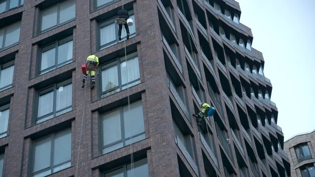 Rope access workers suspended on modern brick high rise corner for exterior window cleaning and maintenance beside geometric facade lines and large windows under overcast daylight