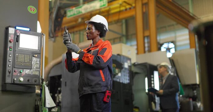 African woman machinist inspecting a cutting tool at a CNC machine control panel in a factory
