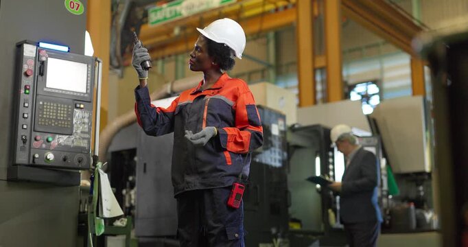 African woman machinist inspecting a cutting tool at a CNC machine control panel in a factory
