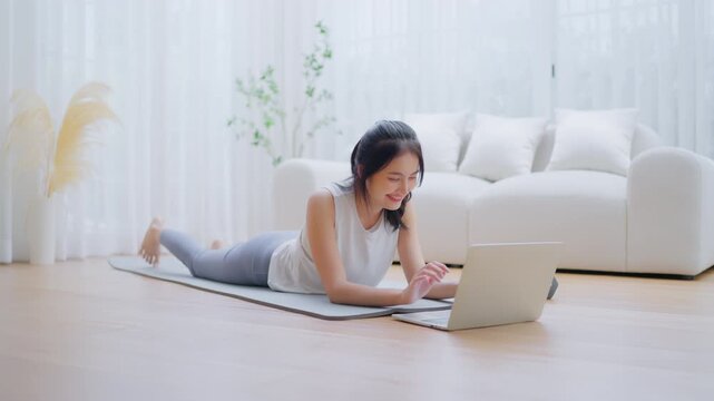 Young Asian woman lying on the floor on yoga mat in front of laptop and training at home. Beautiful smiling woman doing stretching exercise while communicates with a trainer online before workout