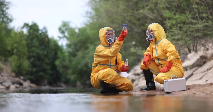 Two environmental technicians in yellow protective suits examine water samples beside a waterway.
