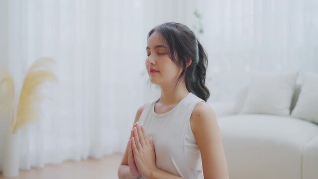 Happy young Asian woman practicing yoga and meditation at home sitting on floor in living room in lotus position and relaxing with closed eyes. Mindful meditation and wellbeing concept