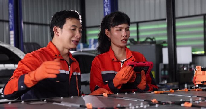 Two automotive technicians discuss an electric vehicle battery inspection and repair plan beside an exposed battery pack. A handheld diagnostic tablet supports their discussion, highlighting teamwork