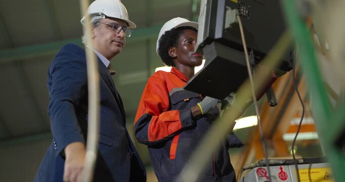 A senior manager in a suit and hard hat stands with a Black female machine operator at a CNC machine control panel, discussing settings and checking production on the factory floor.
