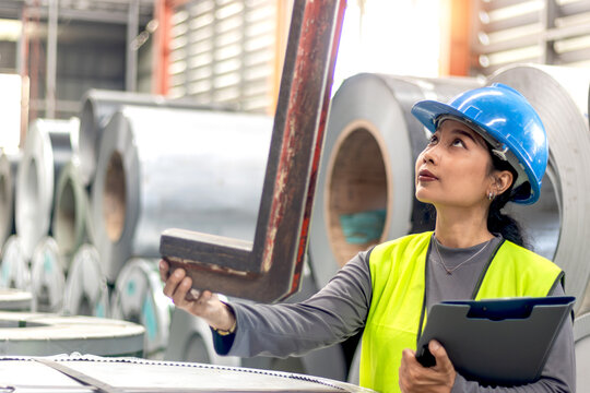 Asian woman worker with helmet and safety vest hold document folder, inspect machine for lifting metal pipes in industrial factory. Female engineer inspector check quality of product at working site.