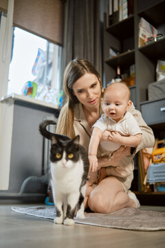 Mother holds her baby in her arms as they watch their cat move through their living room. Sun shines through window, illuminating cozy space filled with books and toys.