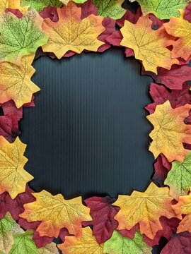 autumn leaves frame. A top-down view of colorful artificial maple leaves arranged in a border frame around a dark textured surface providing copy space for text.