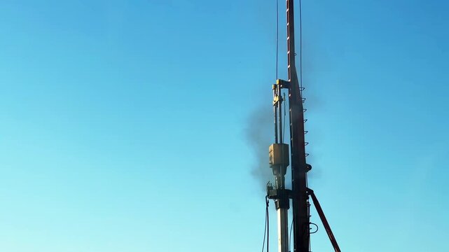 Close-up view of a large construction drilling rig or pile driving machine operating against a clear blue sky. Heavy industrial equipment used for foundation work