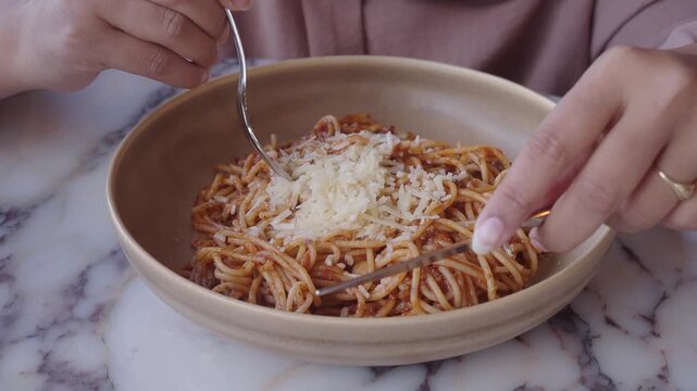 Woman using fork and knife to eat spaghetti with bolognese sauce and grated cheese in bowl.