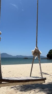 A wooden swing hangs from ropes on a sandy beach. The ocean is calm with distant mountains under a clear blue sky. Ideal for relaxation and summer vibes.